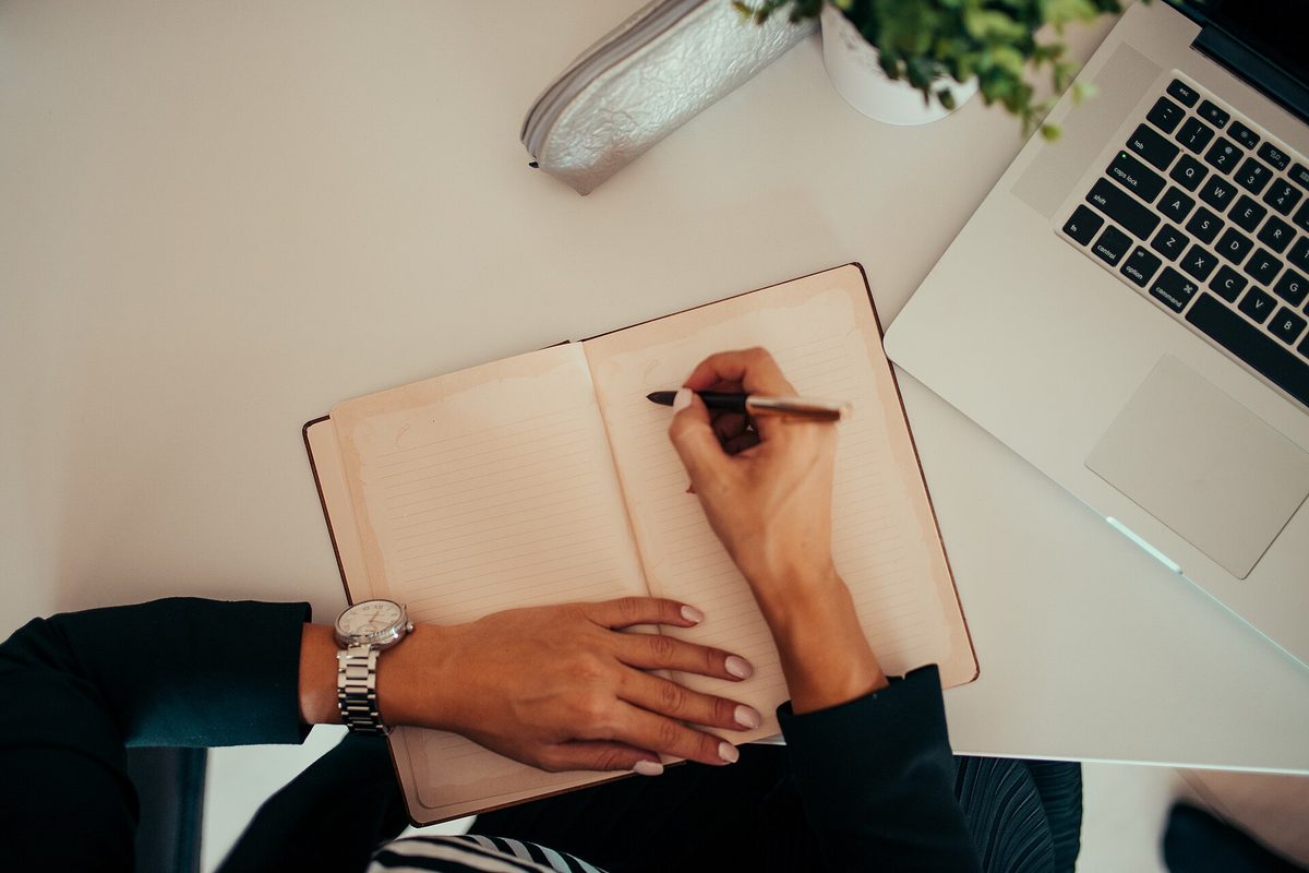 Person writing in notebook while using laptop at a modern workspace
